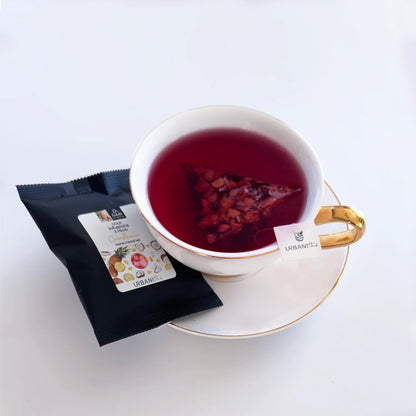 Tasse à thé blanche à anse et soucoupe dorées avec sachet pyramidal Coco-Ananas Urbanithé infusant dans liquide rouge fuchsia, emballage noir Dr Isabelle Coco-Ananas devant, fond blanc
