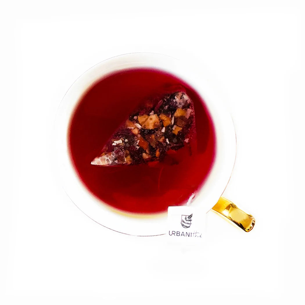 Vue de dessus d’une tasse à anse dorée avec sachet pyramidal Coco-Ananas Urbanithé infusant dans liquide rouge vif, fond blanc