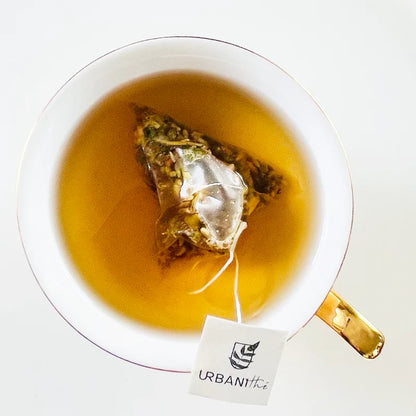Tasse blanche à anse dorée avec sachet Zen Urbanithé infusant, enveloppe individuelle noire Couche-toi avec ça posée devant sur soucoupe blanche