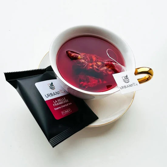 Tisane La Belle Framboise Urbanithé — sachet pyramidal dans tasse à anse dorée avec emballage unitaire noir sur soucoupe blanche