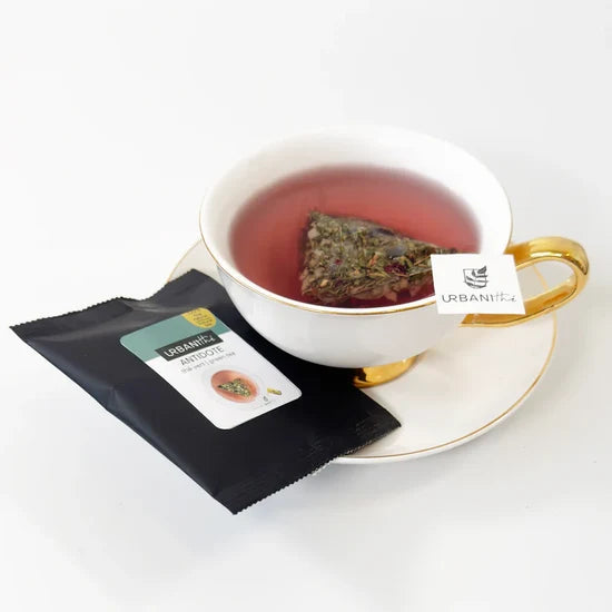 Tasse blanche à anse et soucoupe dorées avec sachet pyramidal Antidote Urbanithé infusant dans liquide rouge ambré, enveloppe individuelle noire Antidote posée devant, fond blanc