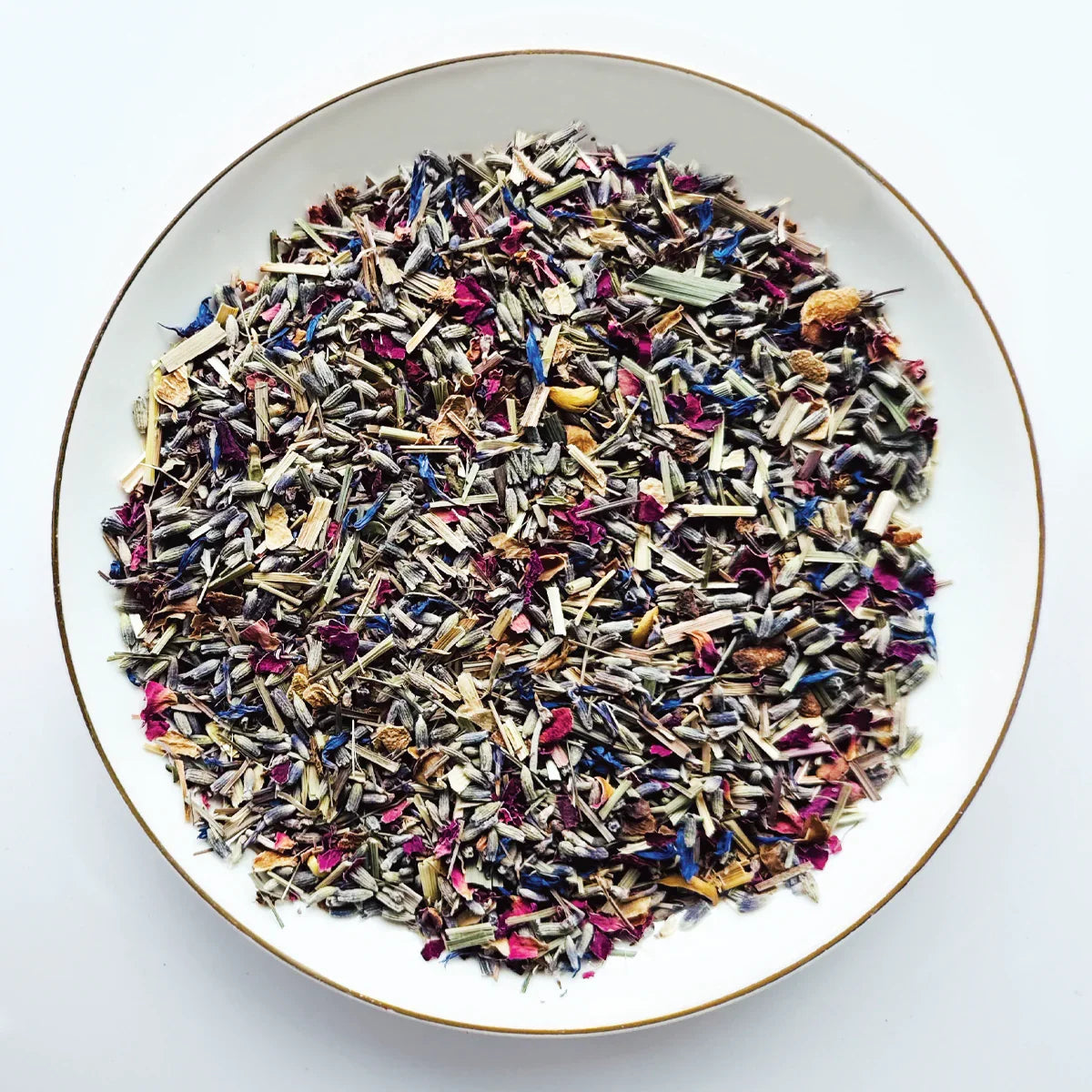Vue de dessus du mélange sec Bleu de Fleurs Urbanithé — lavande bleue, citronnelle, centaurée bleue et pétales de rose dans assiette blanche à rebord doré