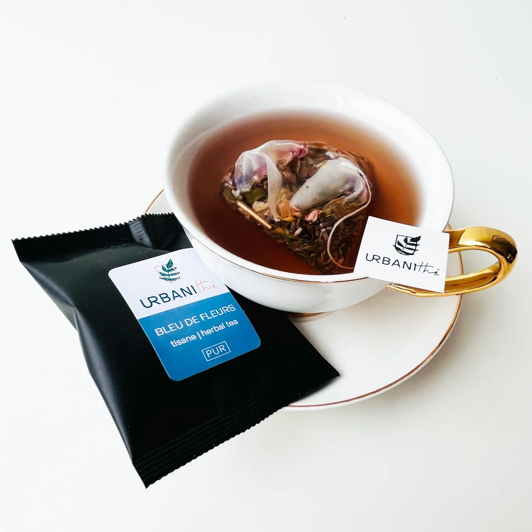 Tasse blanche à anse dorée avec sachet Bleu de Fleurs Urbanithé infusant dans liquide ambré, enveloppe individuelle noire Bleu de Fleurs posée devant, fond blanc