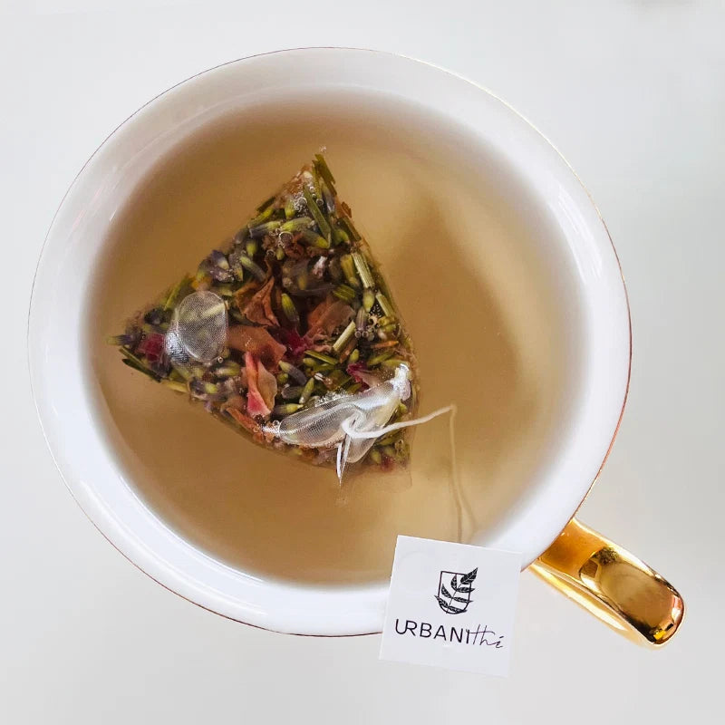 Vue de dessus d'une tasse blanche à anse dorée avec sachet pyramidal Bleu de Fleurs Urbanithé infusant dans liquide doré pâle