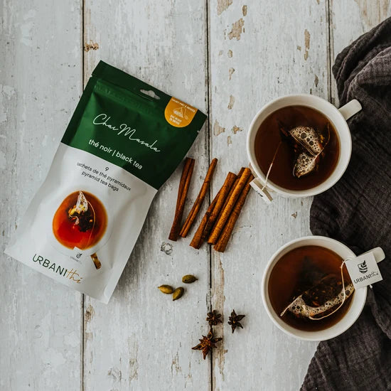 Emballage vert Chai Masala Urbanithé posé sur table de bois blanc avec deux tasses blanches infusant, bâtons de cannelle, cardamome et anis étoilée, serviette grise, ambiance réconfortante