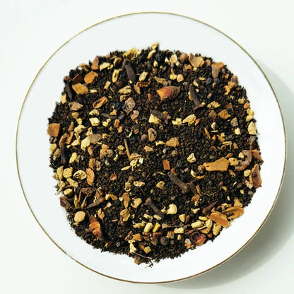 Vue de dessus du mélange sec Chai Masala Urbanithé — thé noir Assam, cannelle, gingembre, cardamome, clous de girofle et épices dans assiette blanche bordée or
