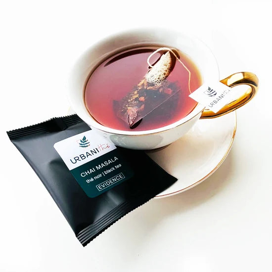 Tasse blanche à anse et soucoupe dorées avec sachet pyramidal Chai Masala Urbanithé infusant, enveloppe individuelle noire Chai Masala Evidence posée devant, fond blanc