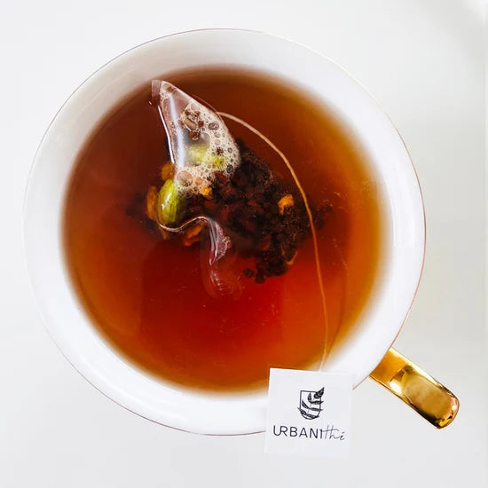 Vue de dessus d’une tasse blanche à anse dorée avec sachet pyramidal Chai Masala Urbanithé infusant dans liquide ambré riche, fond blanc