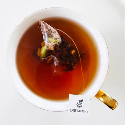 Vue de dessus d’une tasse blanche à anse dorée avec sachet pyramidal Chai Masala Urbanithé infusant dans liquide ambré riche, fond blanc