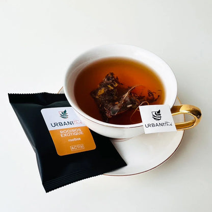 Tasse blanche à anse dorée avec sachet Citron Exotique Urbanithé infusant, enveloppe individuelle noire Rooibos Exotique à côté