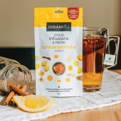 Emballage Citron Exotique Urbanithé jaune et blanc sur table avec tasse en verre infusant, cannelle et tranche de citron en arrière-plan