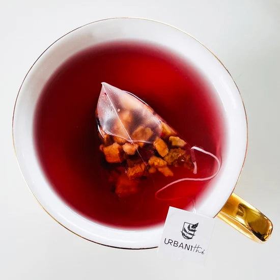 Tasse blanche à anse dorée avec sachet Pomme Canneberge Urbanithé infusant, enveloppe individuelle noire Couverture de Laine Funky posée devant, fond blanc