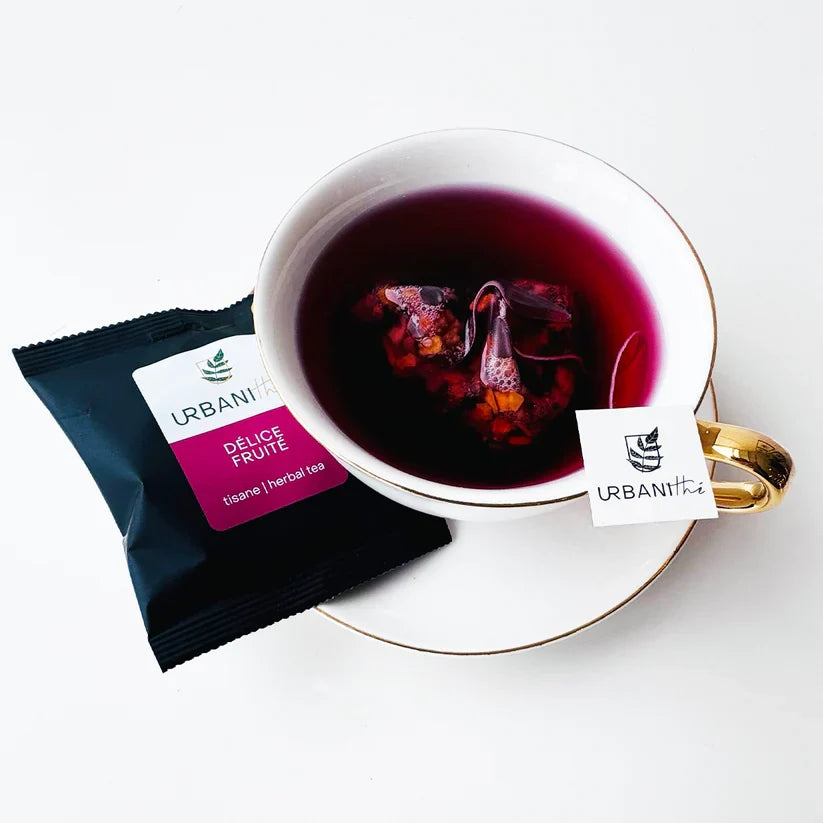 Tasse blanche à anse dorée avec sachet Délice Fruité Urbanithé infusant, enveloppe individuelle noire à côté, fond blanc