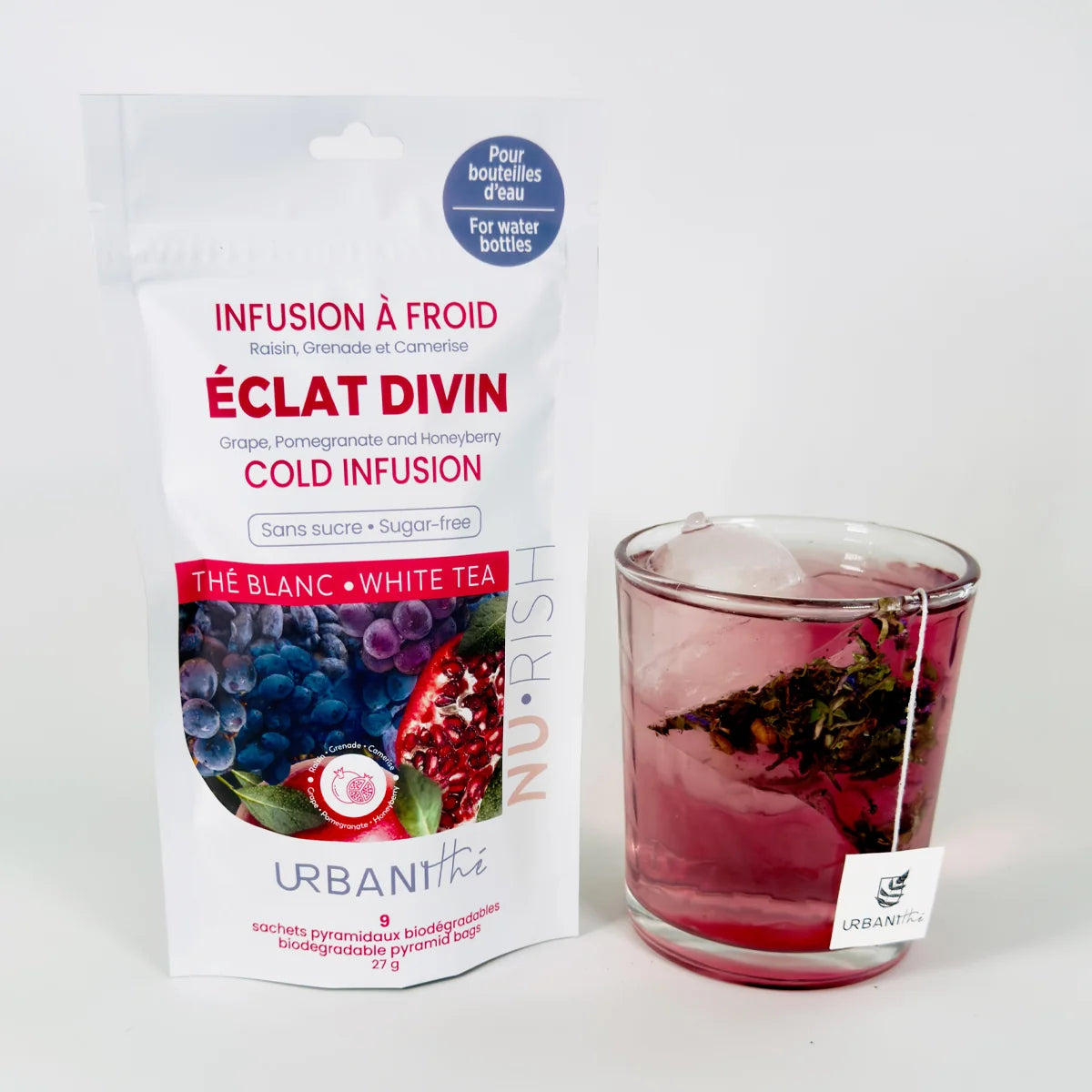 Emballage Éclat Divin Urbanithé x Nu•Rish posé à côté d'un verre carré avec infusion rose-rouge glacée et sachet pyramidal