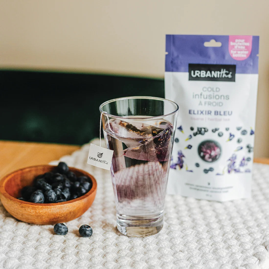 Verre transparent avec infusion Elixir Bleu Urbanithé et sachet pyramidal infusant, bol de bleuets frais et emballage bleu à côté