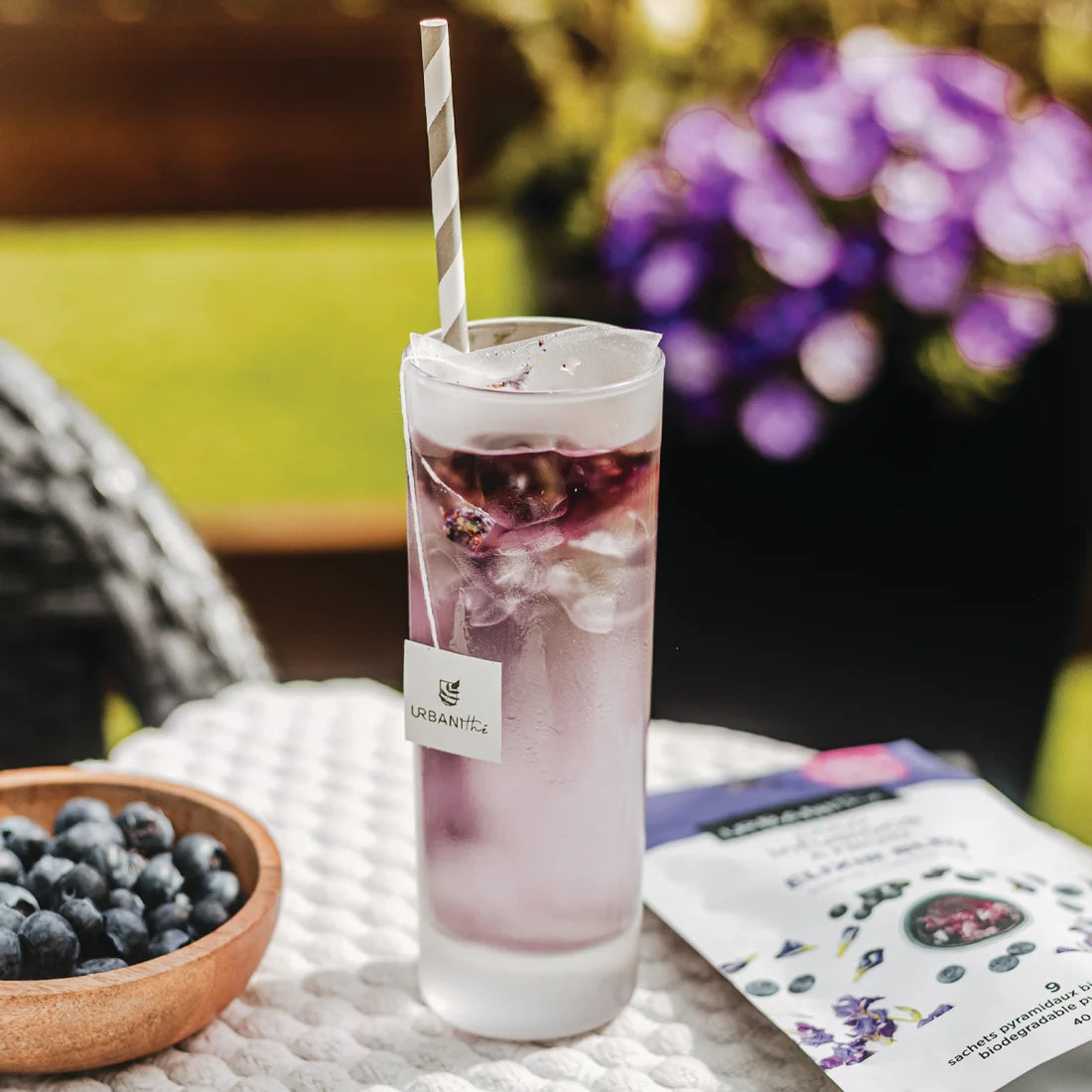 Grand verre glacé avec paille rayée et sachet Elixir Bleu Urbanithé sur table de terrasse, bleuets frais et emballage en arrière-plan