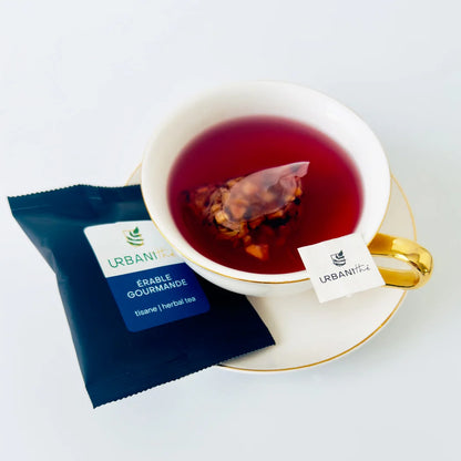 Tasse blanche à anse et soucoupe dorées avec sachet pyramidal Érable Gourmande Urbanithé infusant dans liquide rouge vif, enveloppe individuelle bleue Érable Gourmande posée devant, fond blanc
