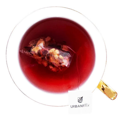 Vue de dessus d’une tasse en verre transparent à anse dorée avec sachet pyramidal Érable Gourmande Urbanithé infusant dans liquide rouge bordeaux profond, fond blanc