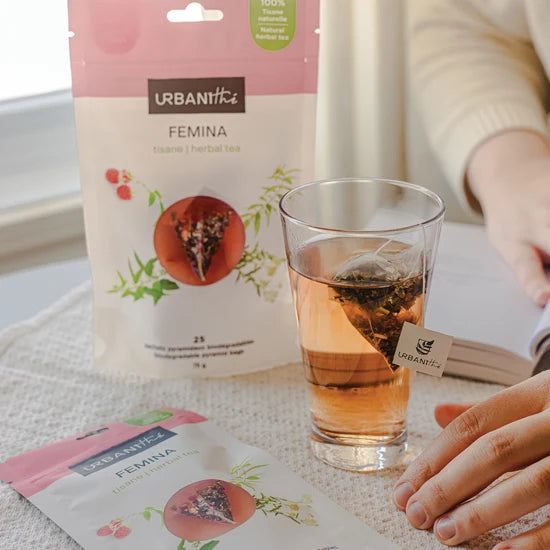Emballage Femina Urbanithé 25 sachets posé derrière un verre en verre avec infusion rose-ambre et sachet pyramidal, enveloppe individuelle Femina devant, ambiance fenêtre lumineuse