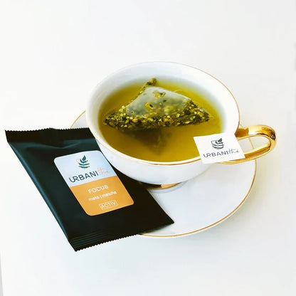 Tasse blanche à anse et soucoupe dorées avec sachet pyramidal Focus Urbanithé infusant dans liquide vert doré, enveloppe individuelle noire Focus posée devant, fond blanc