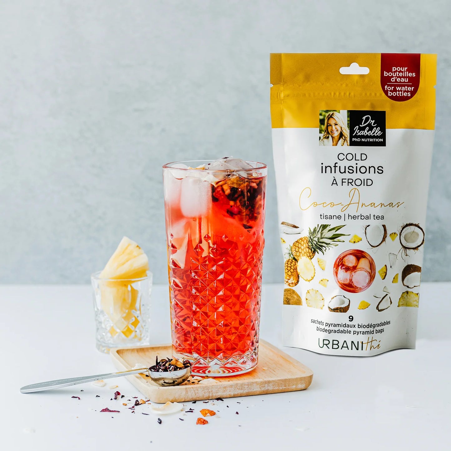 Verre glacé de tisane Coco-Ananas Urbanithé rouge vif avec emballage jaune et blanc, ananas et cuillère à infusion