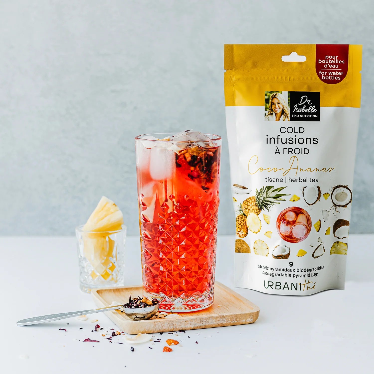 Verre glacé de tisane Coco-Ananas Urbanithé rouge vif avec emballage jaune et blanc, ananas et cuillère à infusion