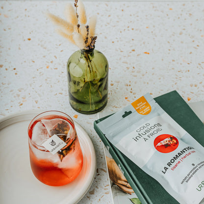 Tasse blanche à anse dorée avec sachet pyramidal La Romantique Urbanithé infusant — liquide rouge profond, emballage noir rose à côté