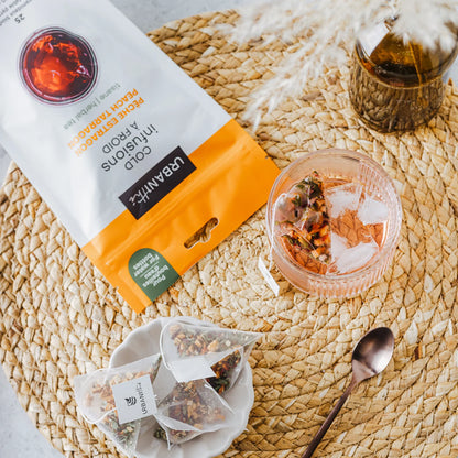 Emballage Pêche Estragon Urbanithé 25 sachets posé derrière une tasse jaune moutarde avec infusion chaude et sachet ouvert sur napperon tressé