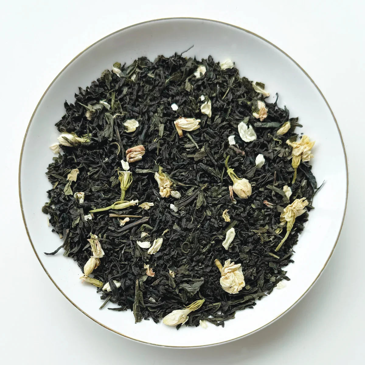 Vue de dessus du mélange sec Pêche Jasmin Urbanithé — thé vert, pétales de jasmin blancs dans assiette blanche à rebord doré