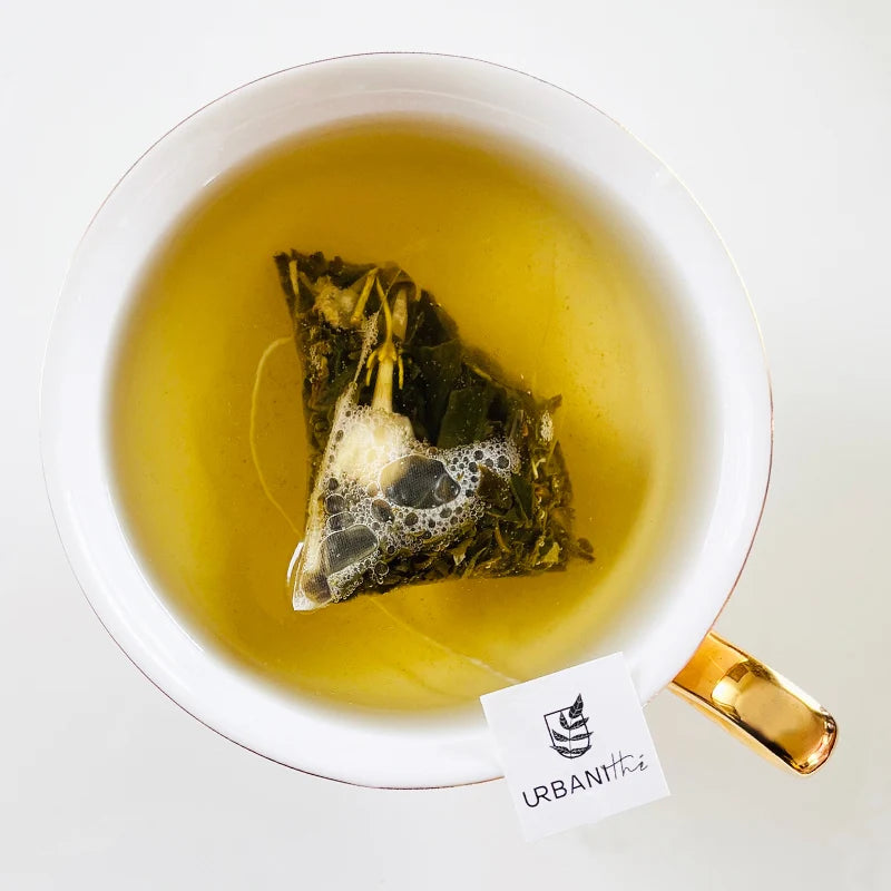 Vue de dessus d’une tasse blanche à anse dorée avec sachet pyramidal Pêche Jasmin Urbanithé infusant dans liquide doré