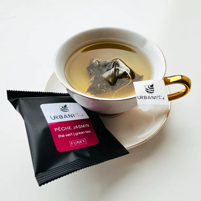 Tasse blanche à anse dorée avec sachet Pêche Jasmin Urbanithé infusant, enveloppe individuelle noire à côté, fond blanc