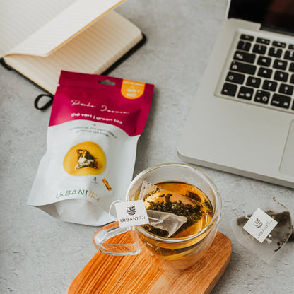 Verre en cristal avec sachet Pêche Jasmin Urbanithé sur planche de bois, emballage framboise et ordinateur portable en arrière-plan
