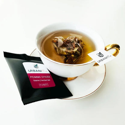 Tasse blanche à anse et soucoupe dorées avec sachet pyramidal Pomme Épicée Urbanithé infusant dans liquide ambré, enveloppe individuelle noire Pomme Épicée posée devant, fond blanc