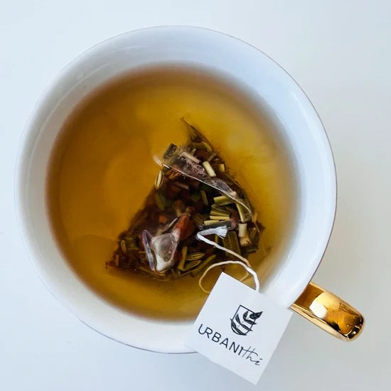 Vue de dessus d’une tasse blanche à anse dorée avec sachet pyramidal Pomme Épicée Urbanithé infusant dans liquide ambré doré, fond blanc