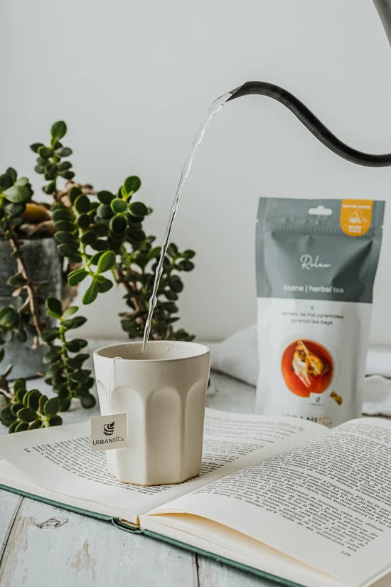 Bouilloire versant eau chaude dans tasse beige avec sachet Relax Urbanithé, emballage 9 sachets posé sur livre ouvert, plante succulente en arrière-plan