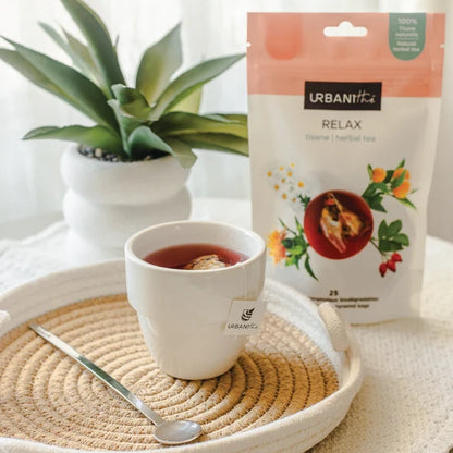 Vue carrée de dessus d'une tasse blanche à anse dorée avec sachet pyramidal Relax Urbanithé infusant dans liquide rouge ambré, enveloppe noire Relax devant, fond blanc