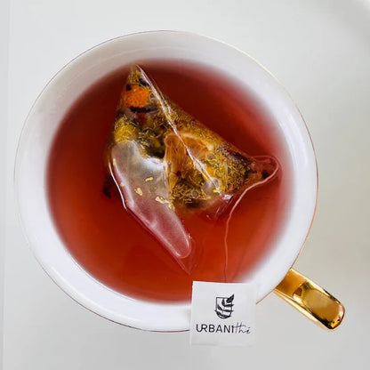 Tasse blanche à anse et soucoupe dorées avec sachet pyramidal Relax Urbanithé infusant dans liquide rouge profond, enveloppe individuelle bleue Relax posée devant, fond blanc