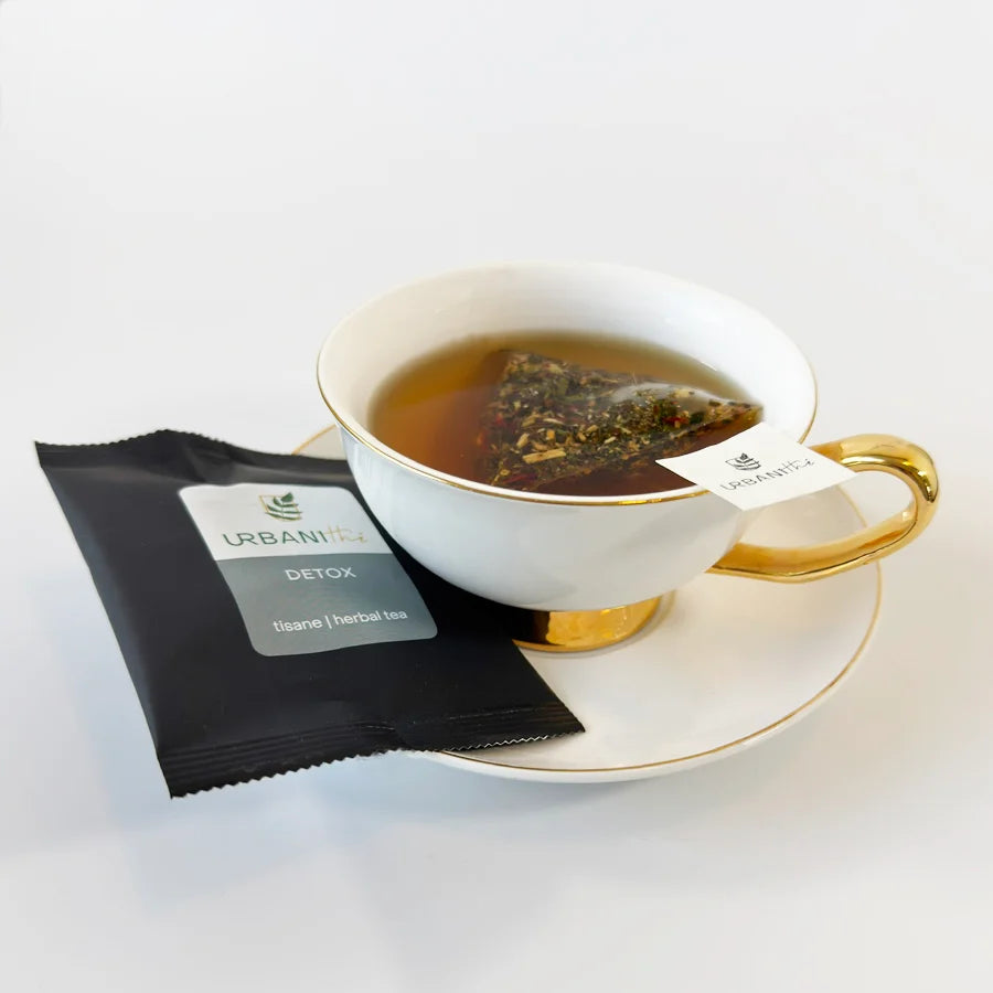 Emballage individuel noir Tisane Detox Urbanithé avec sachet pyramidal infusant dans une tasse blanche à anse dorée sur soucoupe